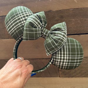 Könnte beinhalten: Ein Haarreif mit zwei runden Ohren, die mit einem dunkel olivgrünen Stoff mit weißem Gittermuster bezogen sind. Eine große, passende Stoffschleife ist mittig zwischen den Ohren platziert und zeigt ebenfalls das olivgrüne und weiße Karomuster.