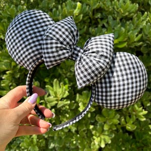 May include: Black and white gingham pattern mouse ear headband with a matching bow. The ears are round and plush, and the bow is centered between them. The headband is black and the background is green foliage.