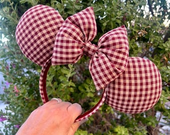 May include: A headband featuring two round, padded ears covered in a red and white gingham fabric. A large, matching red and white gingham fabric bow is centred between the two ears. The headband frame is also wrapped in the same red and white gingham.