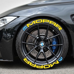 May include: Black car wheel with the word "MOPAR" in yellow lettering on the tire. The wheel has a black rim with a BMW logo in the center and blue brake calipers.