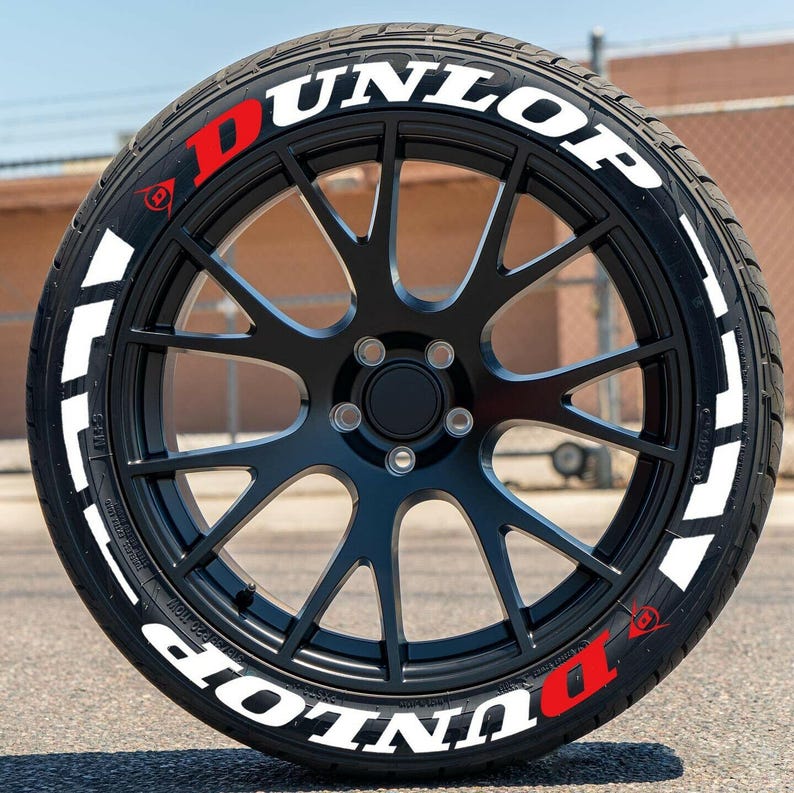 Puede incluir: Un neum&aacute;tico negro con letras blancas que dicen "DUNLOP" alrededor de la banda lateral. El neum&aacute;tico tiene una "D" roja a cada lado.