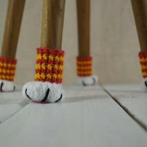 May include: Four wooden chair legs with crocheted paw-shaped covers. The covers are red and yellow striped with white paw pads and black claws.