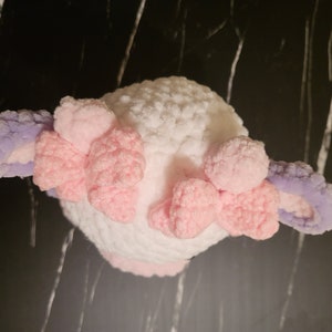 A white crocheted stuffed animal with pink and purple crocheted ears. The animal has a fluffy texture and is shaped like a small animal.  The soft, purple and pink crochet cow toy top-down view, illustrating the top of the head, bows, horns, and texture achievable with this Super Bulky yarn pattern.