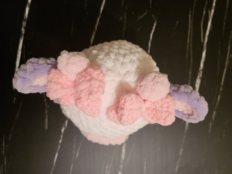 A white crocheted stuffed animal with pink and purple crocheted ears. The animal has a fluffy texture and is shaped like a small animal.  The soft, purple and pink crochet cow toy top-down view, illustrating the top of the head, bows, horns, and texture achievable with this Super Bulky yarn pattern.