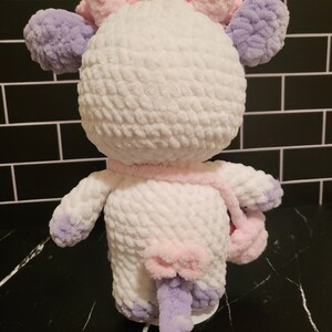 A white crocheted plush toy shaped like a cow with purple and pink accents. The cow has a pink bow on its tail and a removeable pink purse in its hand. The cow is standing on a black surface.