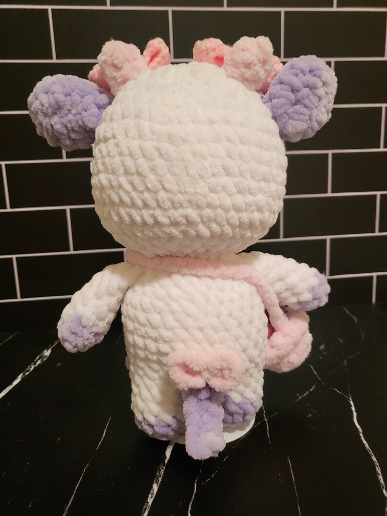 A white crocheted plush toy shaped like a cow with purple and pink accents. The cow has a pink bow on its tail and a removeable pink purse in its hand. The cow is standing on a black surface.
