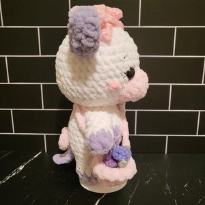 May include: A white crocheted plush toy with purple and pink accents. The toy has a pink bow on its head and is holding a pink and purple basket.