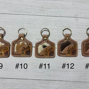 K&ouml;nnte beinhalten: F&uuml;nf Leder-Schl&uuml;sselanh&auml;nger mit Metallringen. Jeder Schl&uuml;sselanh&auml;nger hat eine rechteckige Form mit einem Kuhfell-Einsatz und einem bemalten Sonnenblumen-Design. Die Schl&uuml;sselanh&auml;nger sind mit #9, #10, #11, #12 und #13 nummeriert.