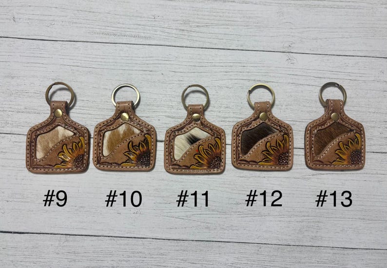 K&ouml;nnte beinhalten: F&uuml;nf Leder-Schl&uuml;sselanh&auml;nger mit Metallringen. Jeder Schl&uuml;sselanh&auml;nger hat eine rechteckige Form mit einem Kuhfell-Einsatz und einem bemalten Sonnenblumen-Design. Die Schl&uuml;sselanh&auml;nger sind mit #9, #10, #11, #12 und #13 nummeriert.