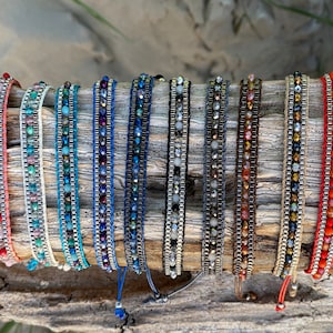 May include: A collection of ten colorful beaded bracelets with different colored cords and beads. The bracelets are displayed on a piece of driftwood.