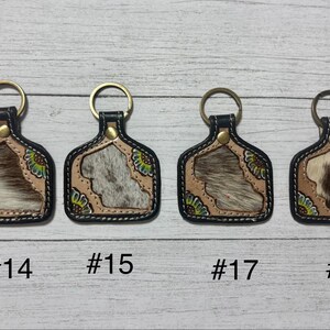K&ouml;nnte beinhalten: Vier Schl&uuml;sselanh&auml;nger mit Leder- und Kuhfell-Design. Jeder Schl&uuml;sselanh&auml;nger hat einen goldenen Ring und einen schwarzen Rand. Die Kuhfellmuster variieren in der Farbe, mit Beige-, Braun- und Wei&szlig;t&ouml;nen. Die Schl&uuml;sselanh&auml;nger sind mit den Nummern #14, #15, #17 und #18 versehen.