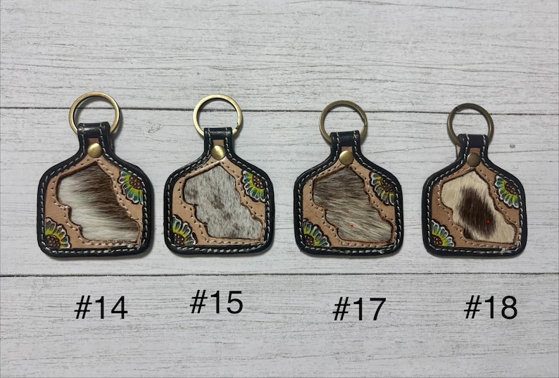 K&ouml;nnte beinhalten: Vier Schl&uuml;sselanh&auml;nger mit Leder- und Kuhfell-Design. Jeder Schl&uuml;sselanh&auml;nger hat einen goldenen Ring und einen schwarzen Rand. Die Kuhfellmuster variieren in der Farbe, mit Beige-, Braun- und Wei&szlig;t&ouml;nen. Die Schl&uuml;sselanh&auml;nger sind mit den Nummern #14, #15, #17 und #18 versehen.