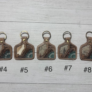 K&ouml;nnte beinhalten: F&uuml;nf Leder-Schl&uuml;sselanh&auml;nger mit Metallringen. Jeder Schl&uuml;sselanh&auml;nger hat einen braunen Lederrahmen mit floralem Design und einen Abschnitt aus Kuhfell. Die Schl&uuml;sselanh&auml;nger sind mit #4, #5, #6, #7 und #8 nummeriert.
