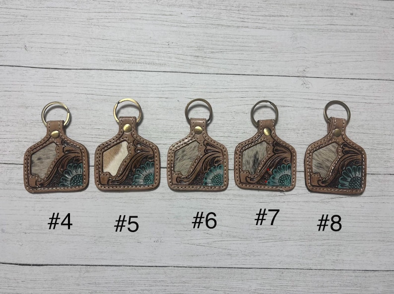 K&ouml;nnte beinhalten: F&uuml;nf Leder-Schl&uuml;sselanh&auml;nger mit Metallringen. Jeder Schl&uuml;sselanh&auml;nger hat einen braunen Lederrahmen mit floralem Design und einen Abschnitt aus Kuhfell. Die Schl&uuml;sselanh&auml;nger sind mit #4, #5, #6, #7 und #8 nummeriert.
