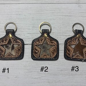 K&ouml;nnte beinhalten: Drei Leder-Schl&uuml;sselanh&auml;nger mit Stern-Design. Jeder Schl&uuml;sselanh&auml;nger hat einen schwarzen Rand, einen goldfarbenen Ring und einen dekorativen Stern in der Mitte. Die Sterne haben unterschiedliche Texturen und Farben, mit den Nummern #1, #2 und #3 darunter.