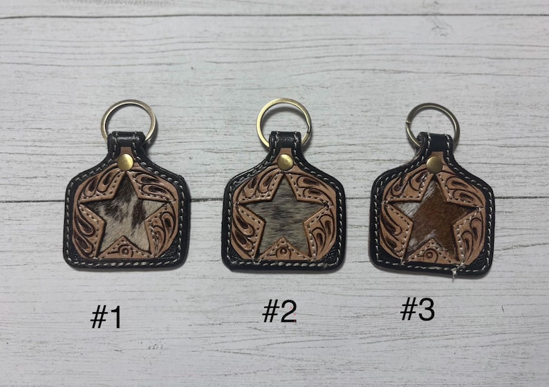 K&ouml;nnte beinhalten: Drei Leder-Schl&uuml;sselanh&auml;nger mit Stern-Design. Jeder Schl&uuml;sselanh&auml;nger hat einen schwarzen Rand, einen goldfarbenen Ring und einen dekorativen Stern in der Mitte. Die Sterne haben unterschiedliche Texturen und Farben, mit den Nummern #1, #2 und #3 darunter.
