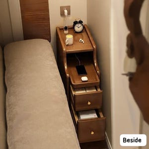 May include: A wooden nightstand with a built-in charging station, two drawers, and a top shelf. The top shelf holds a black alarm clock, a white mug, and a small lamp. A smartphone is charging on the middle shelf. The nightstand is next to a bed.