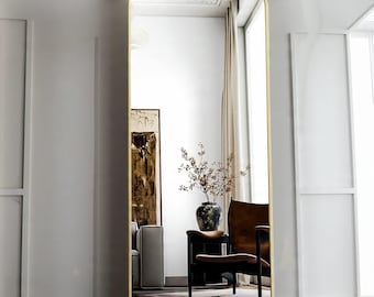 May include: A tall, arched-top mirror with a gold-colored frame. The mirror reflects a room with white walls, a sofa, a chair, and a vase of flowers. The word "Gold" is in the bottom right corner.