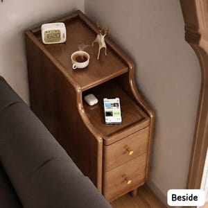 May include: A wooden bedside table with two drawers and a curved design. The top shelf holds a digital clock, a coffee cup, and a deer figurine. A smartphone and wireless earbuds rest on the middle shelf. The table has gold-colored drawer pulls.