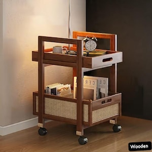 May include: A wooden, two-tiered rolling cart with a dark brown finish. The top shelf holds a mug, glasses, and a clock. The lower shelf has a woven rattan panel and holds books and magazines. The cart has black wheels.