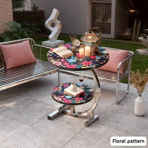 May include: A two-tiered side table with a floral pattern on the tabletop. The table has a chrome frame and a curved support. The top tier has a lamp, candles, and a book. The bottom tier has a book and a small vase. The text "Floral pattern" is at the bottom.