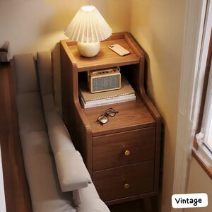 May include: A wooden nightstand with two drawers and a top shelf. A small lamp with a pleated shade sits on top, along with a smartphone and a small radio. Books and eyeglasses are also visible. The nightstand has gold-colored knobs.