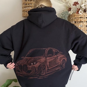 Pode incluir: Uma camisola preta com capuz apresentando uma grande estampa gráfica em tons de castanho de um carro desportivo nas costas. O design mostra o veículo numa vista frontal de três quartos, destacando a grelha, os faróis e as rodas características.