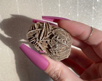 May include: A brown desert rose crystal with a rosette shape. The crystal is held in a hand with pink nail polish.