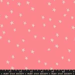 May include: A coral pink fabric featuring a repeating pattern of small, off-white hand-drawn stars scattered across the surface. A black ruler at the bottom edge shows measurements in inches, with the text Ruby Star Society printed repeatedly.
