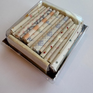 May include: A clear plastic container holds a collection of rolled fabric pieces. The fabric is cream-colored and patterned with floral, geometric, and dotted designs in pastel colors. The container sits on a silver tray.