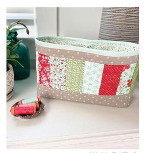 May include: A patchwork fabric basket with the text "Selena Divided Basket". The basket is beige with a polka dot pattern and features panels of red, green, and white floral fabric. Finished size is 7" x 12" x 4".