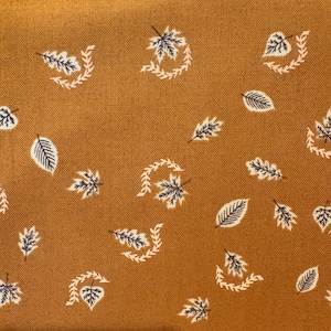 May include: A mustard yellow fabric featuring a repeating pattern of small, dark grey leaves and delicate white vine-like accents. The textile has a visible woven texture, with the leaf motifs scattered across the surface in various orientations.
