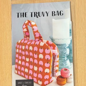 May include: A fabric bag with a handle, featuring a pattern of pink apples on a rust-colored background. The bag is in a clear plastic sleeve. The text "THE TRUVY BAG" is visible. The bag is next to spools of thread and sewing clips.