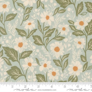 May include: A light blue fabric featuring a repeating pattern of white daisies with orange centers and green leaves. A white ruler is visible at the bottom edge, showing a scale of 7 inches across the width of the textile print.