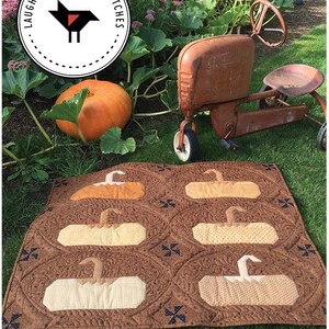 May include: A brown quilt with pumpkin appliqués, a large orange pumpkin, and a vintage toy tractor are displayed on green grass. The quilt features six pumpkin shapes in various shades of tan and cream. The wall hanging is 36" x 30".
