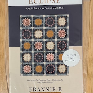 May include: A quilt pattern titled "Eclipse" by Frannie B Quilt Co. The pattern features a quilt design with repeating square blocks in shades of blue, pink, and gold. Finished size is 58" x 72".