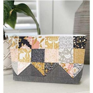May include: A patchwork Wilma Pouch, a mini charm project, with a gray base and a zipper closure. The pouch is made of various patterned fabrics in pink, gold, black, and gray. Finished size is 8" x 11".