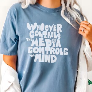 May include: A blue t-shirt with the text "WHOEVER CONTROLS THE MEDIA CONTROLS THE MIND" in white. The shirt is part of the "Comfort Colors" collection and features a round "BLUEJEAN" logo.