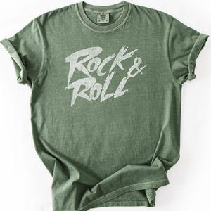 May include: A moss green t-shirt with the words "Rock & Roll" in a distressed white font. The shirt is made of a soft material and has a classic crew neck design. The shirt is a Comfort Colors 1717.