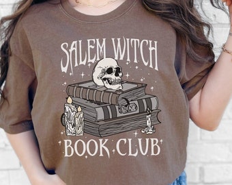 May include: Espresso-colored t-shirt with the text "Salem Witch Book Club" in a gothic font. The design features a skull atop stacked books and lit candles. The shirt is a Comfort Colors 1717.