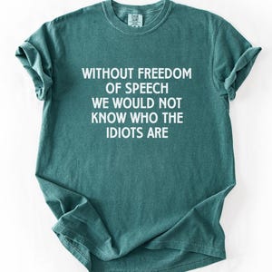 May include: A blue spruce colored short-sleeve t-shirt featuring white text printed on the front that reads: WITHOUT FREEDOM OF SPEECH WE WOULD NOT KNOW WHO THE IDIOTS ARE. The garment has a crew neckline and a standard fit with a textured fabric finish.