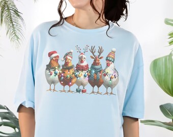 May include: Light blue t-shirt featuring a festive design of six chickens adorned with holiday hats, scarves, and string lights. The shirt has the text "CHAMBRAY" and "COMFORT COLORS 1717". The design is a custom design.