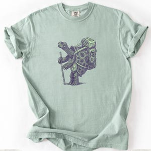 May include: Light green t-shirt featuring a detailed illustration of a turtle with a backpack and walking stick, designed in shades of purple and green. The shirt is a Comfort Colors 1717. The shirt is a light green color.