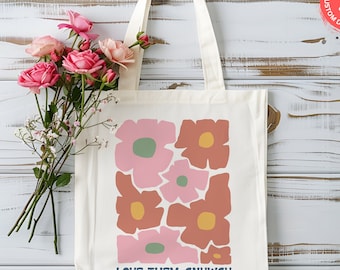 May include: Cream-colored tote bag with a floral design in shades of pink, brown, and yellow. The bag features the text "LOVE THEM ANYWAY" in navy blue. A bouquet of pink roses and delicate white flowers rests beside the bag.
