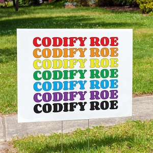 May include: A white sign with the text "CODIFY ROE" repeated seven times in rainbow colors. The sign is on a green lawn.