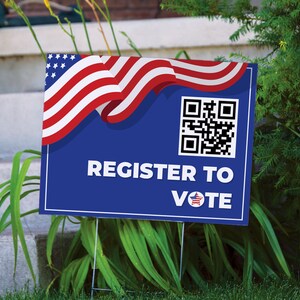 May include: A blue yard sign with the text "REGISTER TO VOTE" and a QR code. The top of the sign features a waving American flag design in red, white, and blue. The sign is placed in a grassy area.