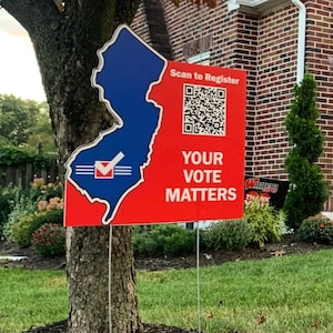 May include: A red and blue sign with the outline of New Jersey. The sign reads "Scan to Register" and "Your Vote Matters." A QR code is also present. The sign is placed in a yard near a tree.