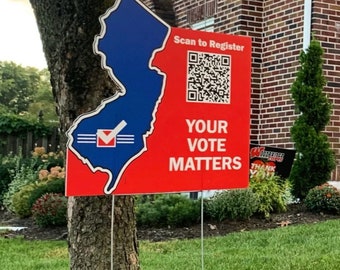 May include: A red and blue sign in the shape of New Jersey, with the text "Scan to Register" and "Your Vote Matters." A QR code is also visible. The sign is placed in a garden near a tree.