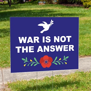 May include: A blue yard sign with the message "WAR IS NOT THE ANSWER" in white text. A white dove carrying an olive branch is at the top, and a red poppy with green and yellow accents is at the bottom. The sign is displayed on a grassy lawn.