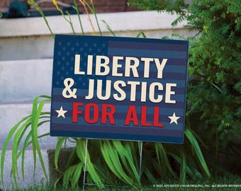 May include: A rectangular sign with the text "LIBERTY & JUSTICE FOR ALL" in white and red against a blue background with a flag design. The sign is supported by metal stakes and is placed in a grassy area.